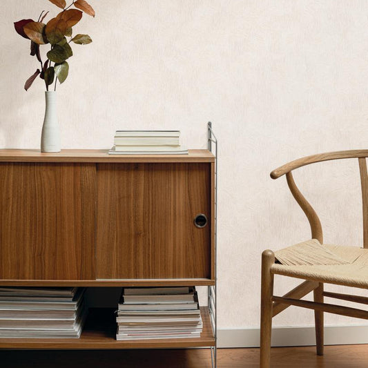 Beige textured wallpaper shown in a styled interior scene with a wooden cabinet, books, a vase with dried leaves, and part of a wooden chair.