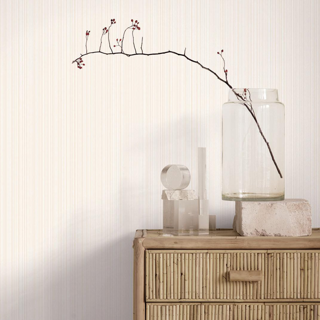 A styled interior scene showing a light wood dresser against a cream wall with subtle vertical texture. On top of the dresser are a clear glass jar containing a single bare branch with small red buds, a clear cylindrical vase, and a small sculptural object.