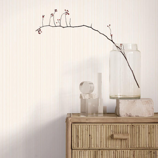 A styled interior scene showing a light wood dresser against a cream wall with subtle vertical texture. On top of the dresser are a clear glass jar containing a single bare branch with small red buds, a clear cylindrical vase, and a small sculptural object.