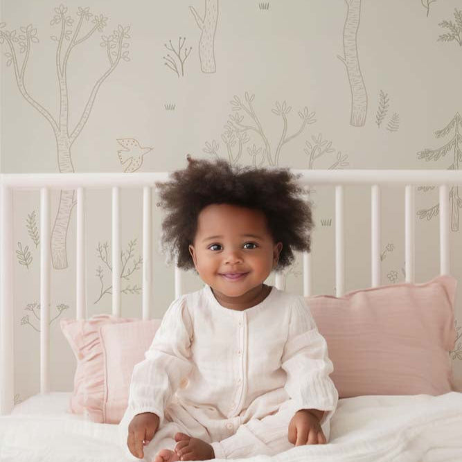Wallpaper Kids Nursery Dubai Peps Wall A young child sitting on a bed with pillows, in front of a wall covered in forest-themed wallpaper with trees and a forest floor pattern.