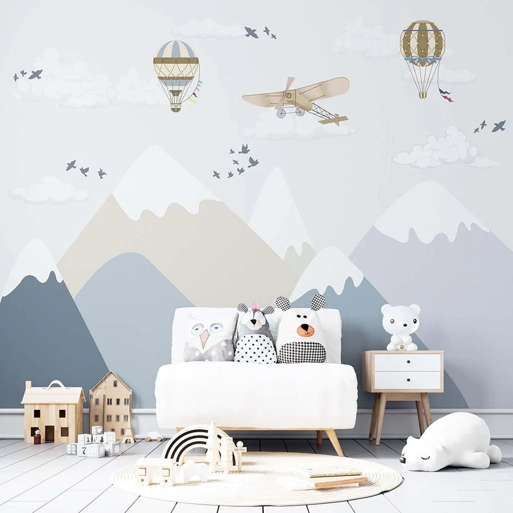 Wallpaper Kids Nursery Dubai Peps Wall A wall mural featuring mountains, clouds, hot air balloons, and airplanes with toy versions of these items on the floor in front of a sofa with children's toys.