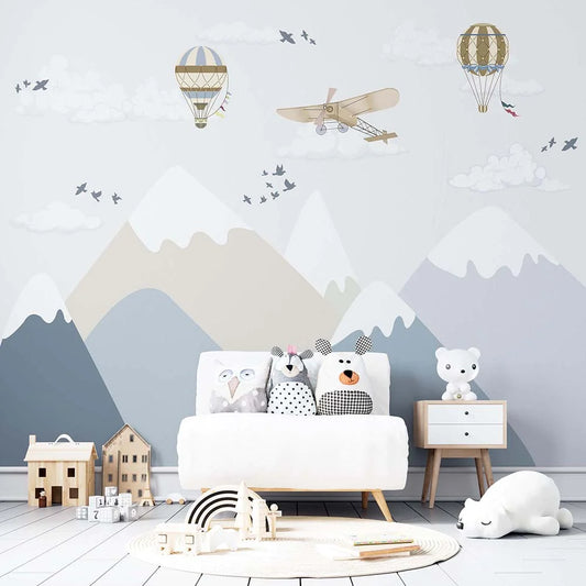 Wallpaper Kids Nursery Dubai Peps Wall A wall mural featuring mountains, clouds, hot air balloons, and airplanes with toy versions of these items on the floor in front of a sofa with children's toys.