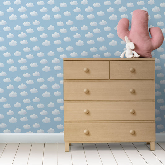 A wooden dresser with a beige finish, featuring a pattern of white clouds on a blue background, accompanied by a pink cactus decal and a small white teddy bear on top.